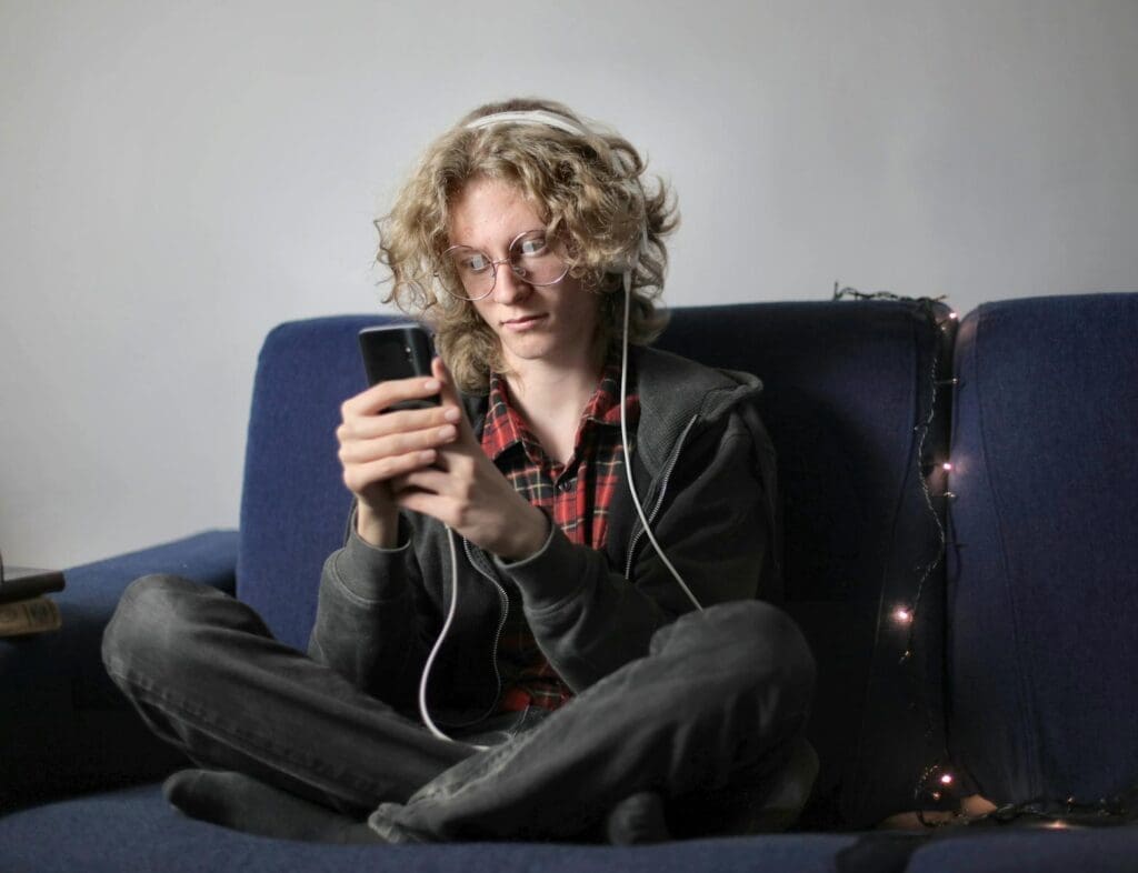 Streaming Audio Trends. Long haired teenager in circle eyeglasses and casual wear sitting on blue soft couch with luminous garland messaging on mobile phone and listening to music on headphones at home